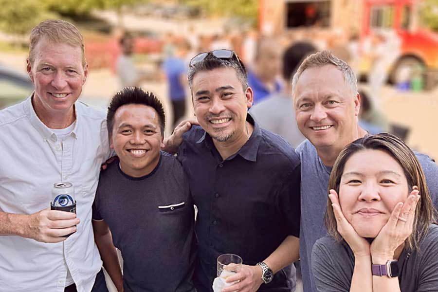Five smiling adults posing closely together outdoors at a social event with blurred people and a food truck in the background.