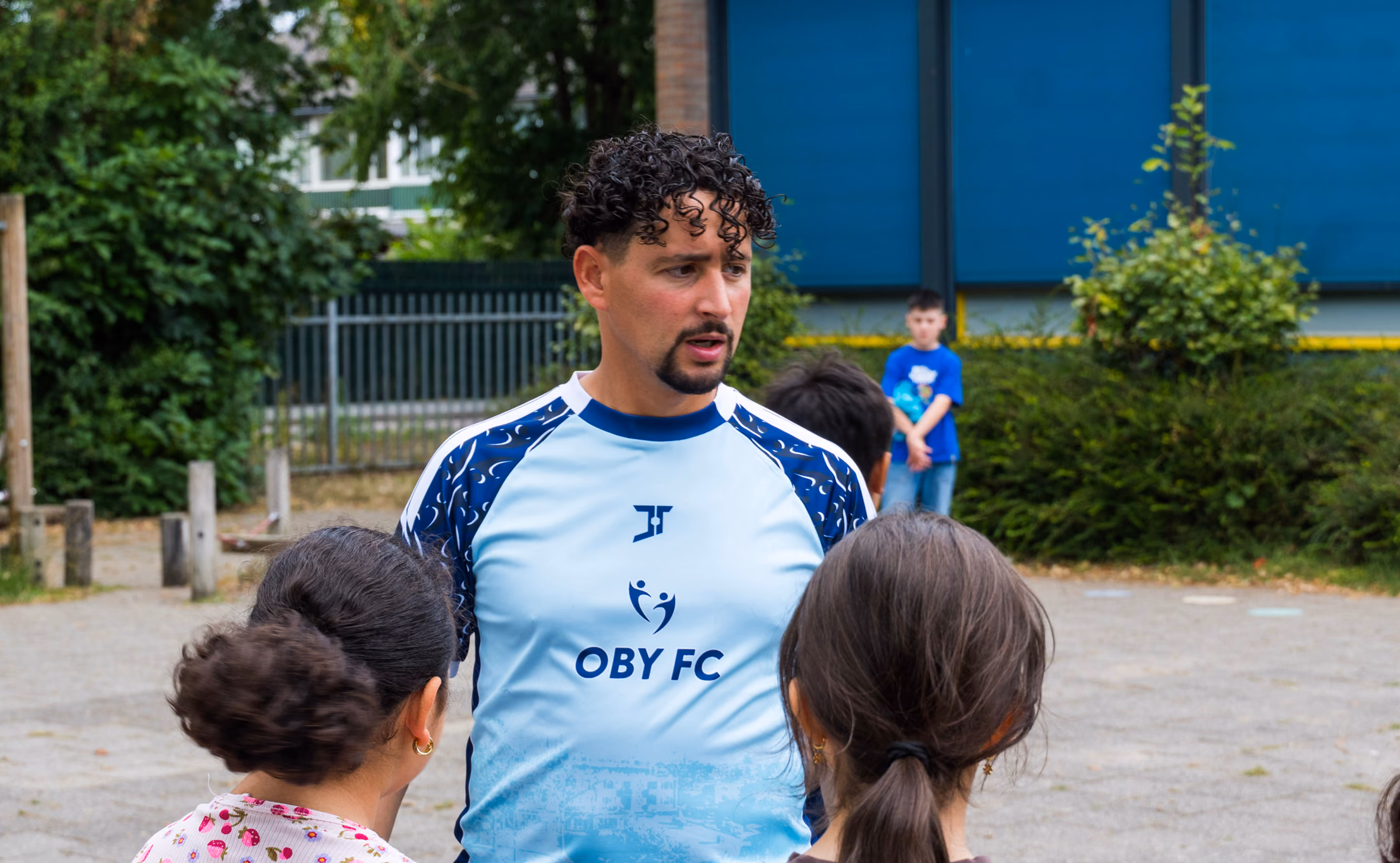 A man with curly hair and a goatee wearing a light blue Oby FC sports shirt talking to children outdoors with trees and a building in the background.