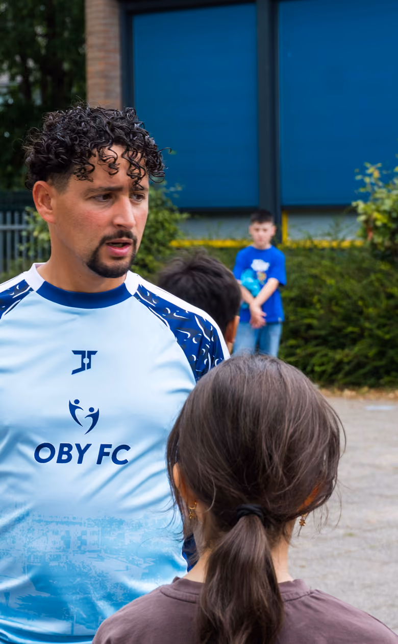 A man with curly hair and a goatee wearing a light blue Oby FC sports shirt talking to children outdoors with trees and a building in the background.