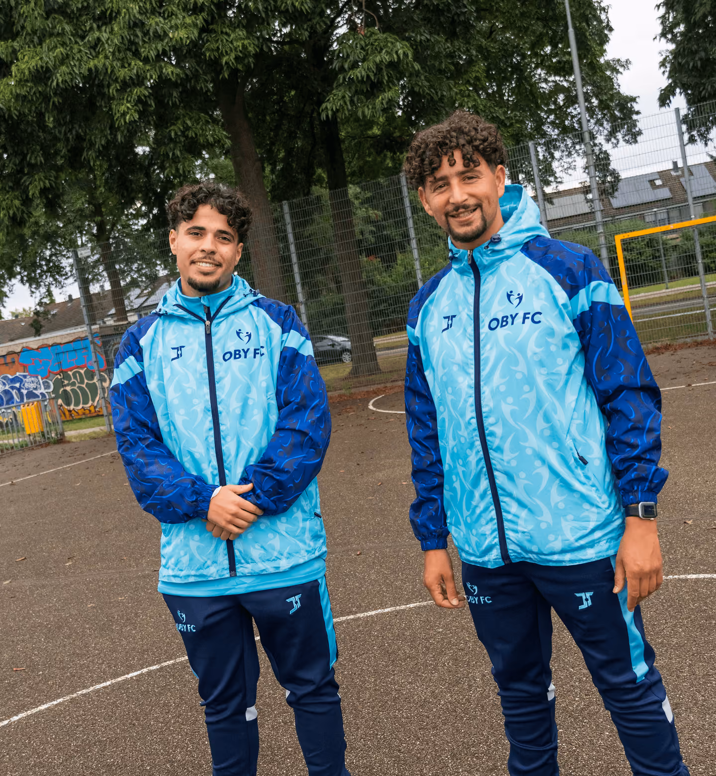 Two men with curly hair wearing matching light blue and navy blue OBY FC jackets standing outdoors near a fenced sports court with trees and graffiti in the background.