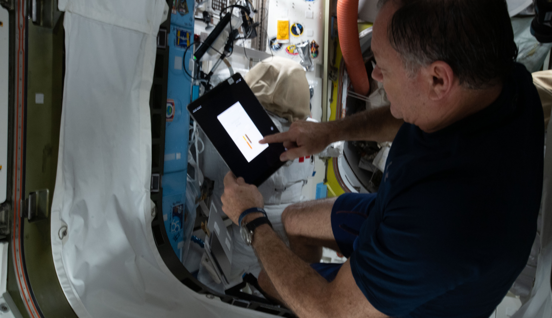 Astronaut inside a spacecraft using a digital tablet mounted in front of various equipment panels.