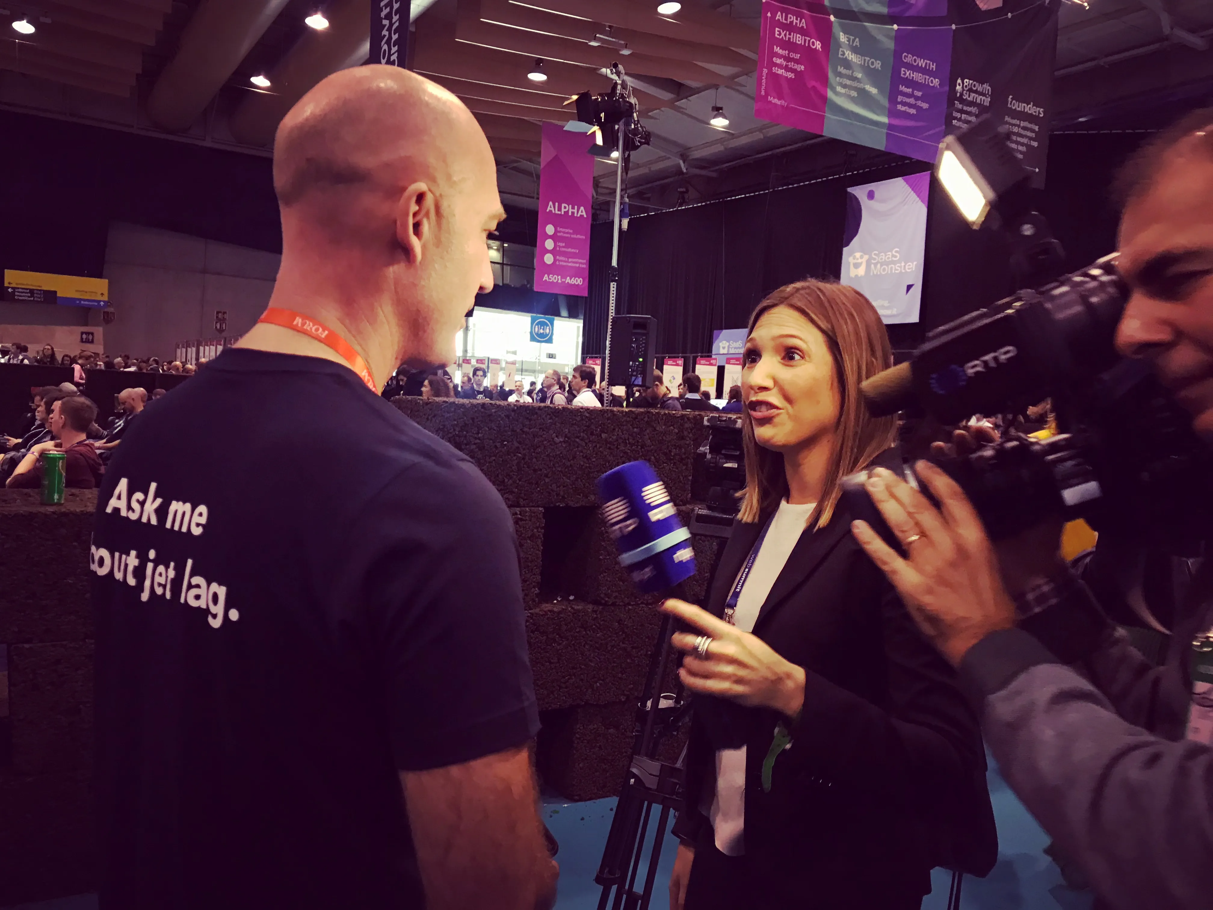 Woman interviewing a bald man wearing a shirt that says 'Ask me about jet lag' while a cameraman films them at an indoor event.