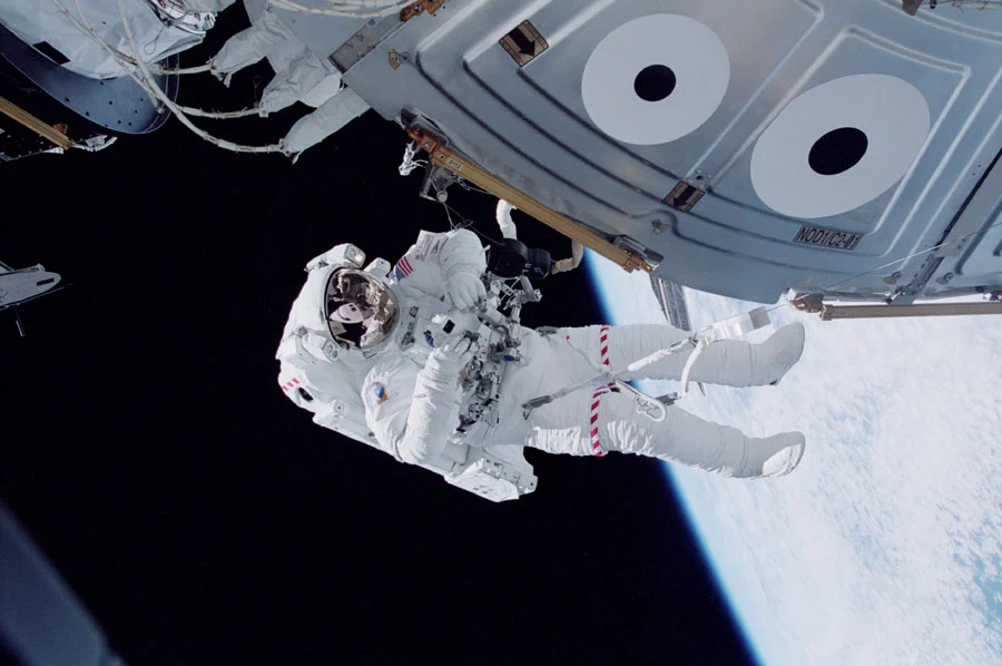 Astronaut in white spacesuit performing a spacewalk outside a spacecraft with Earth and sunlight in the background.