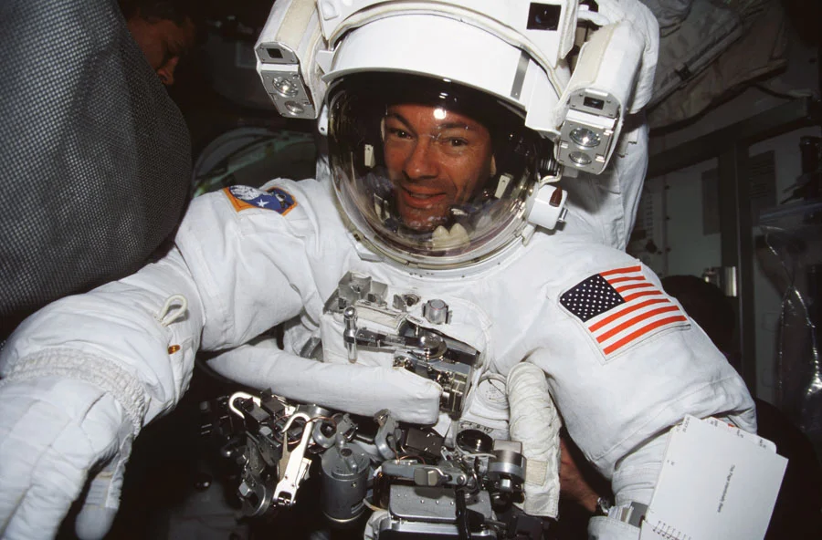 Smiling astronaut in a white space suit with an American flag patch, inside a spacecraft.