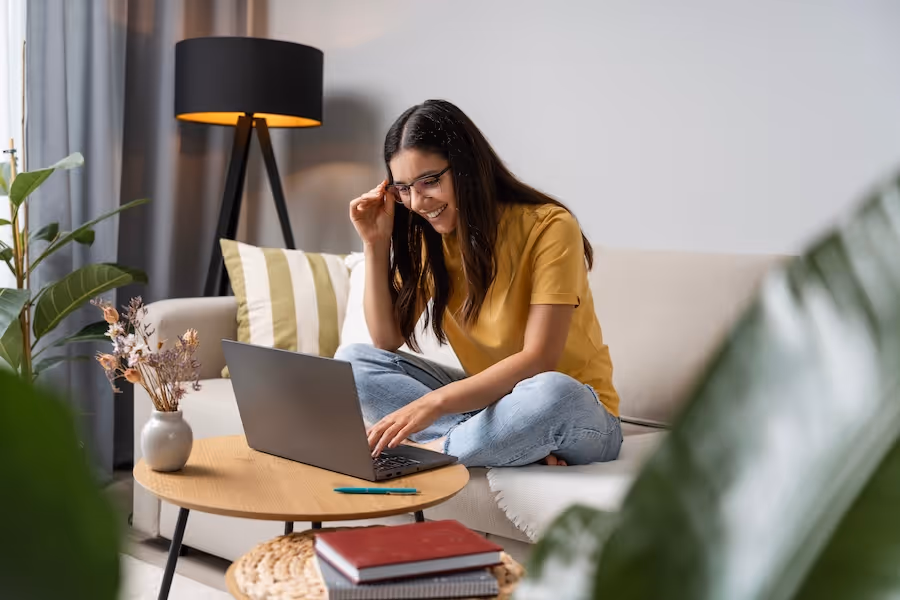Young person with curly hair focused on a laptop while sitting at a wooden table with an open notebook.
