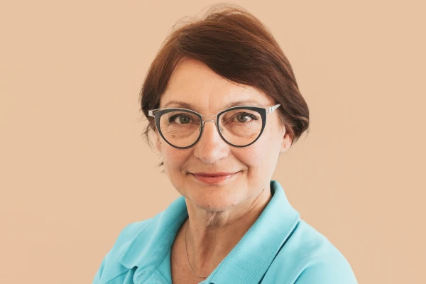 Porträt einer lächelnden älteren Frau mit kurzen braunen Haaren und Brille vor beigem Hintergrund.