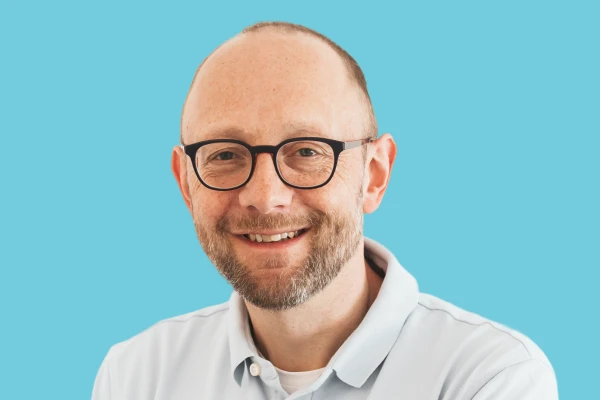 Lächelnder Mann mit Brille, kurzem Bart und Hemd vor blauem Hintergrund.