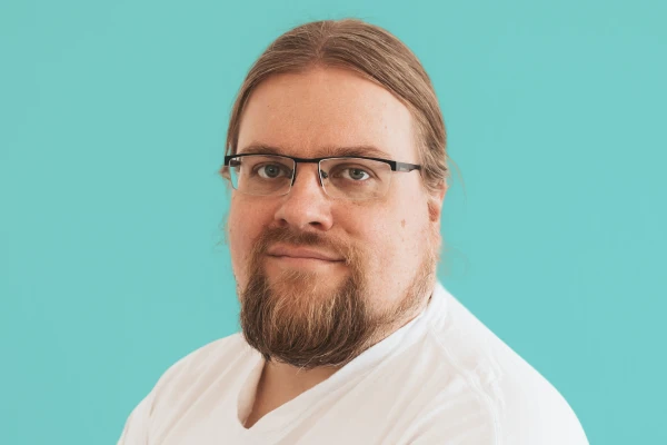 Porträt eines Mannes mit Brille, braunem langem Haar zurückgebunden und Vollbart vor hellblauem Hintergrund.