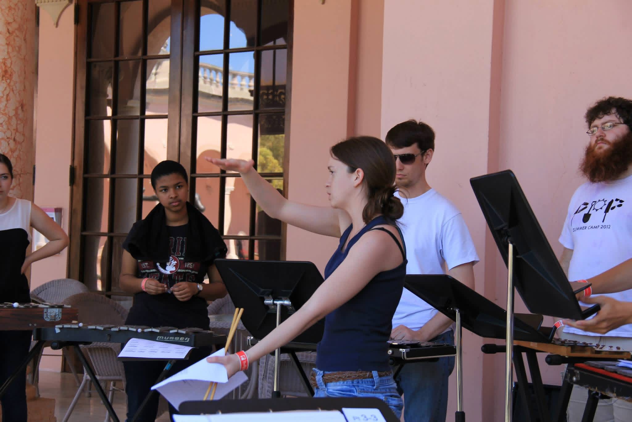 Photograph of Marissa instructing a group of musicians playing mallet percussion instruments outdoors near a building with large windows.