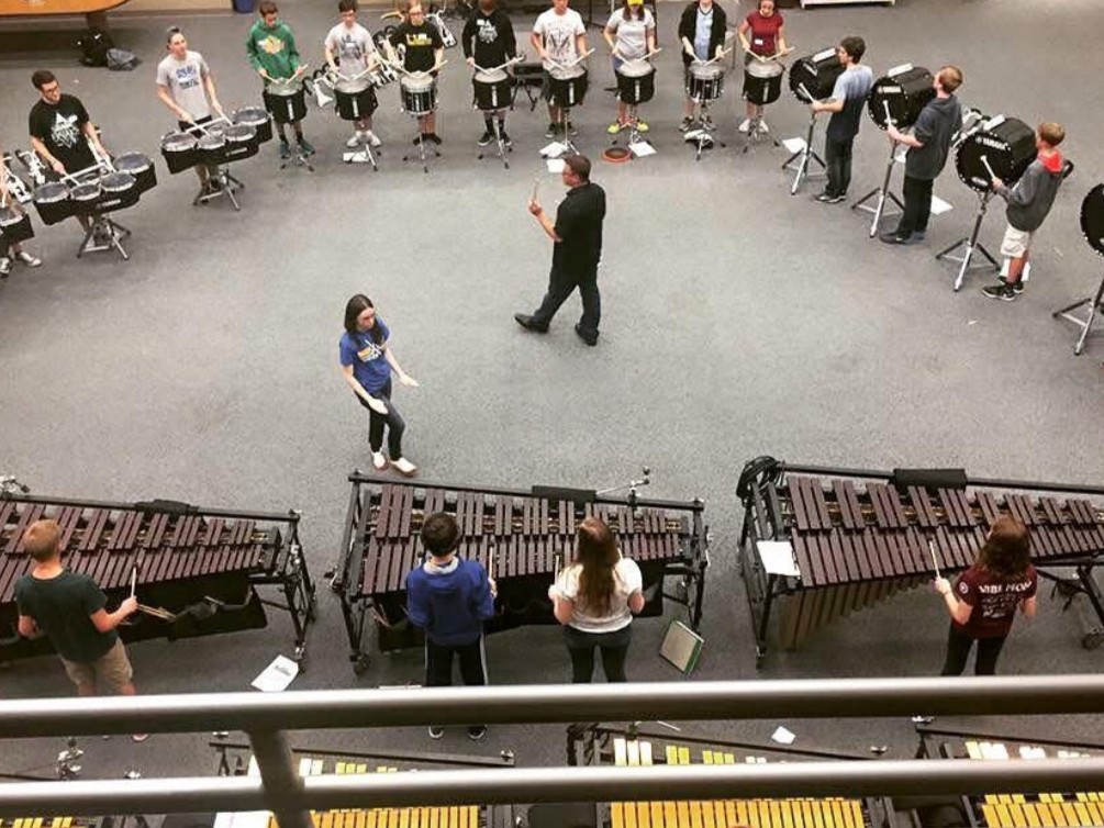 Overhead view of a percussion ensemble with students playing marimbas and drums arranged in a semicircle, with Marissa and another instructor in the center.
