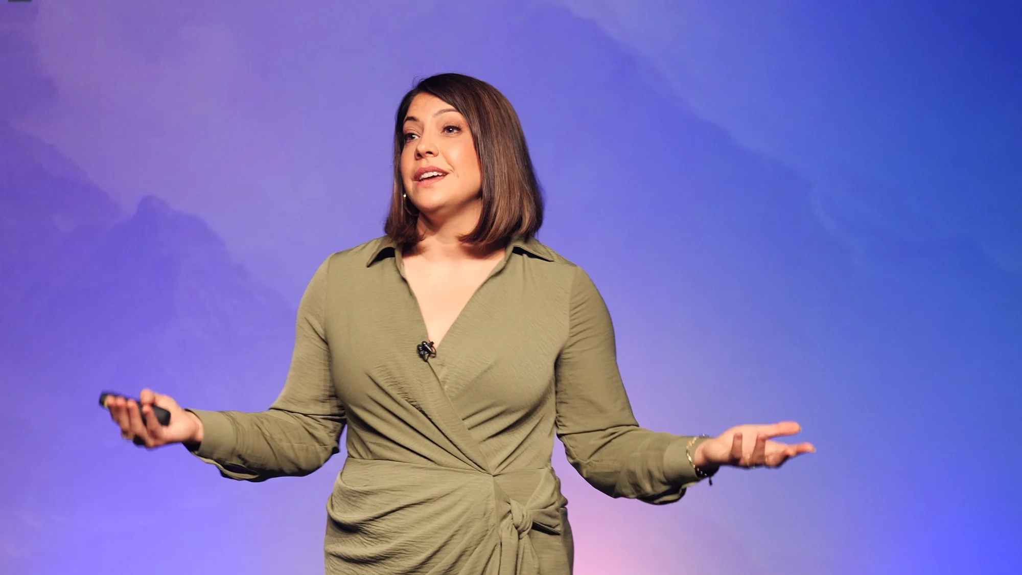Woman with shoulder-length brown hair speaking on stage with arms outstretched against a blue-purple gradient background.