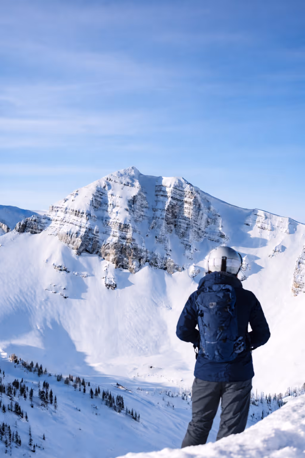 Person in winter gear with a blue backpack overlooking snow-covered mountain under a clear blue sky.