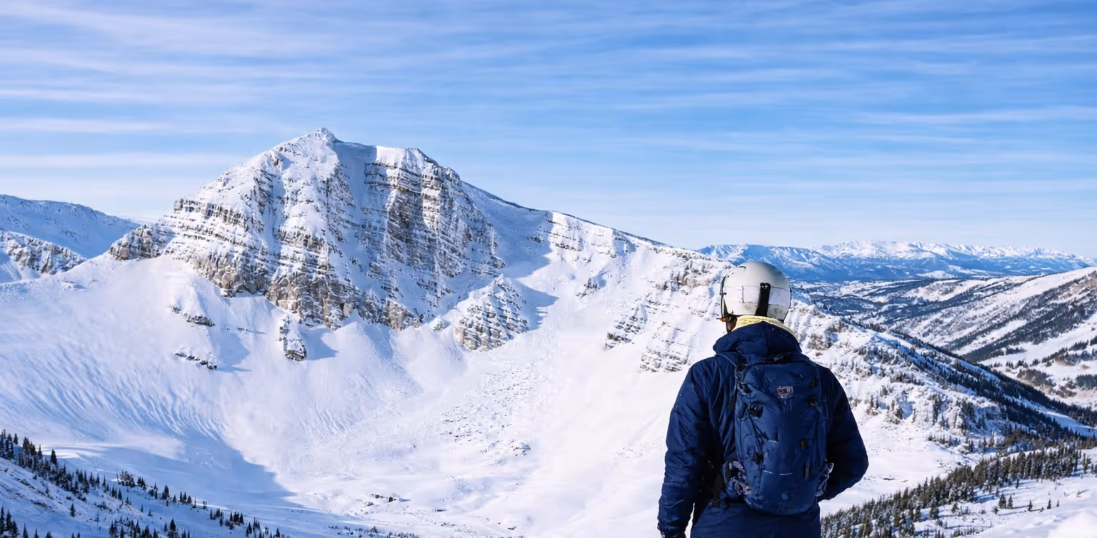 Person wearing a helmet and blue jacket looking at a large snow-covered mountain under a clear blue sky.