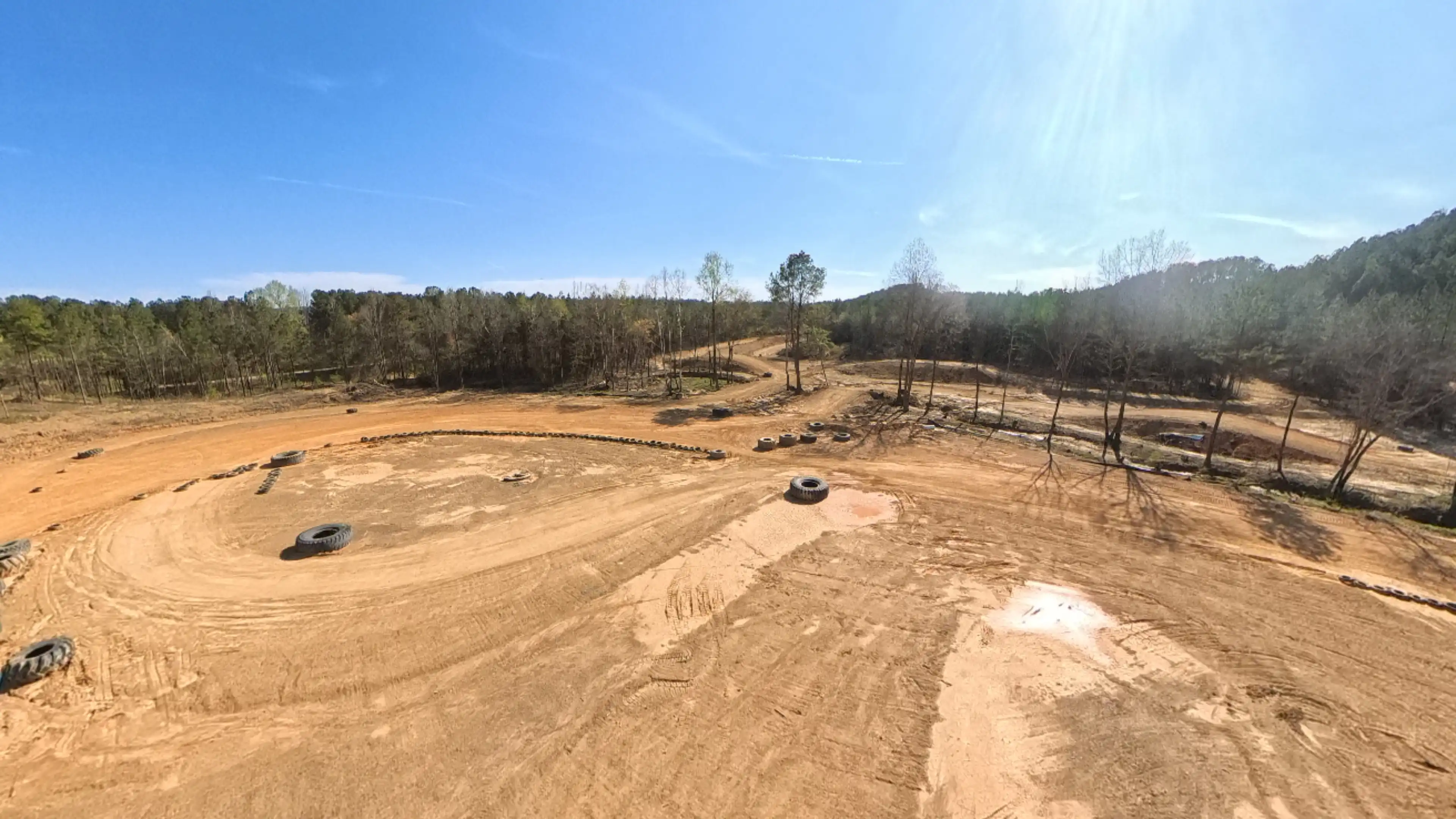 Wide view of a dirt off-road track with scattered tires used as barriers, surrounded by trees under a clear blue sky.
