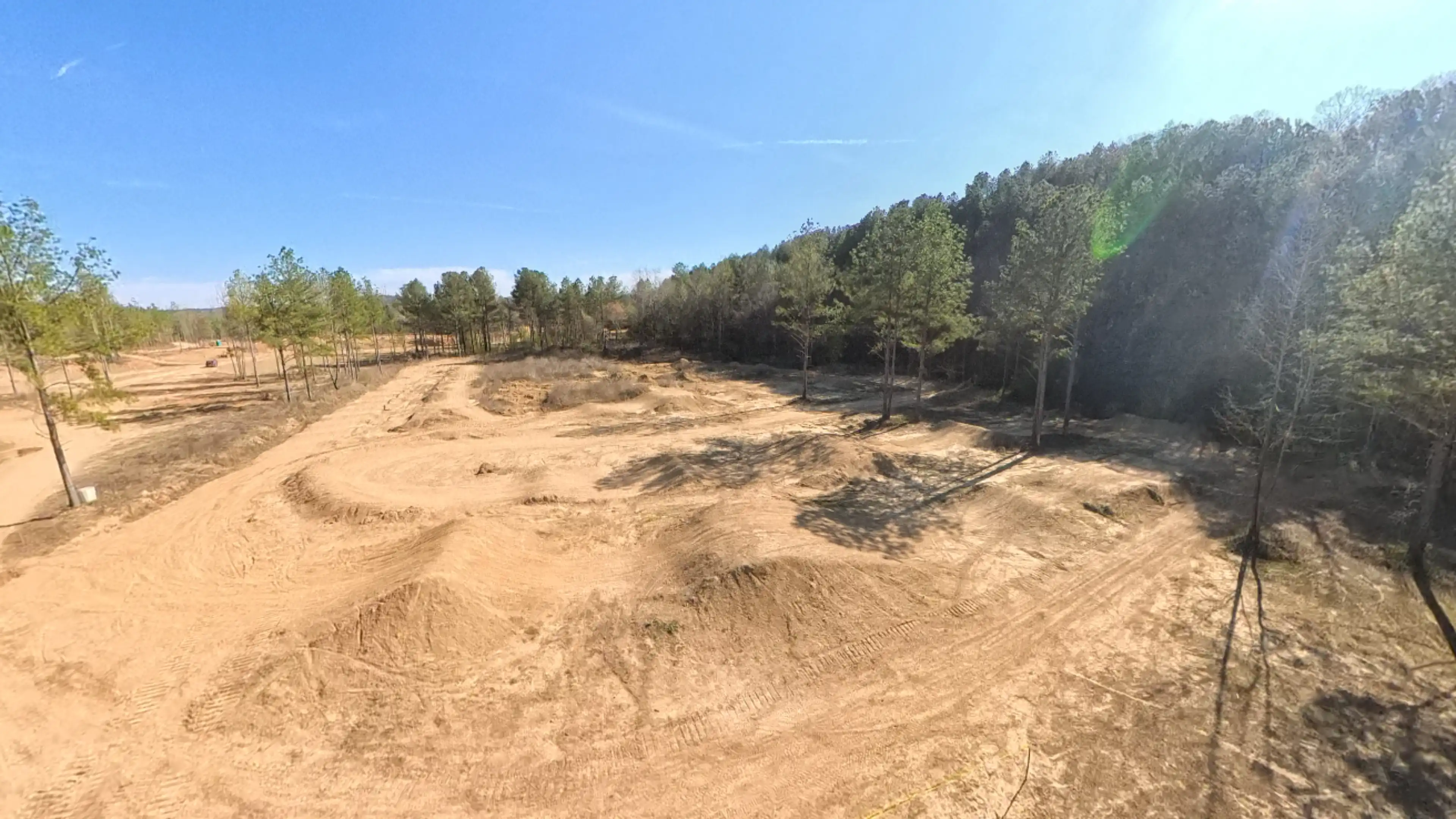 Cleared dirt terrain with tire tracks and small hills under a blue sky, bordered by a forest of pine trees.