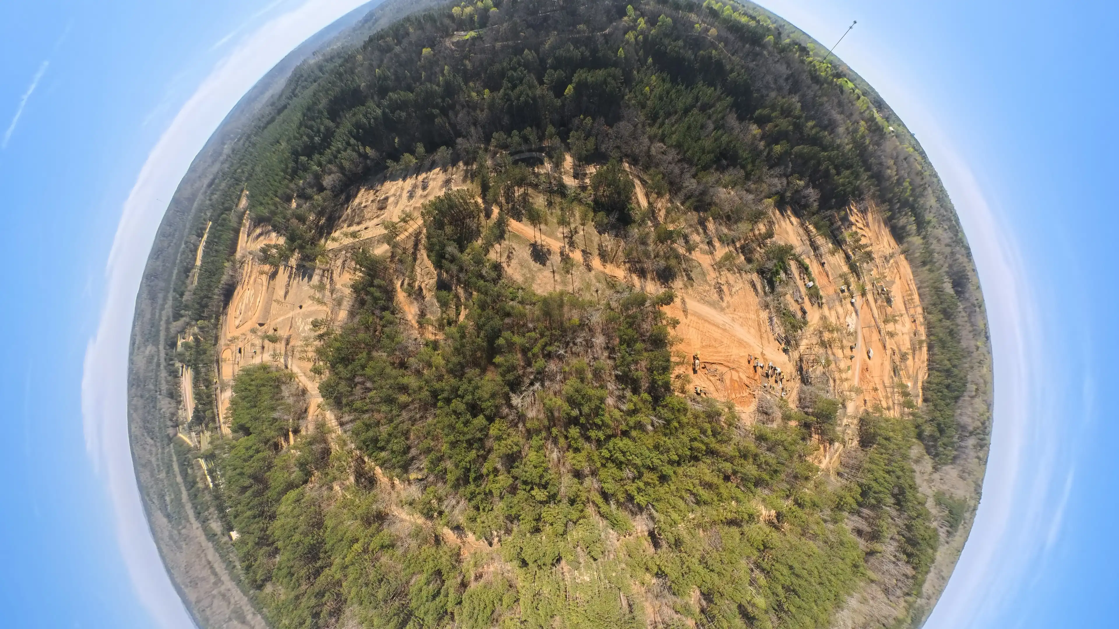 A wide aerial fisheye view showing a circular area of forest surrounding a clearing with dirt roads and some construction vehicles.