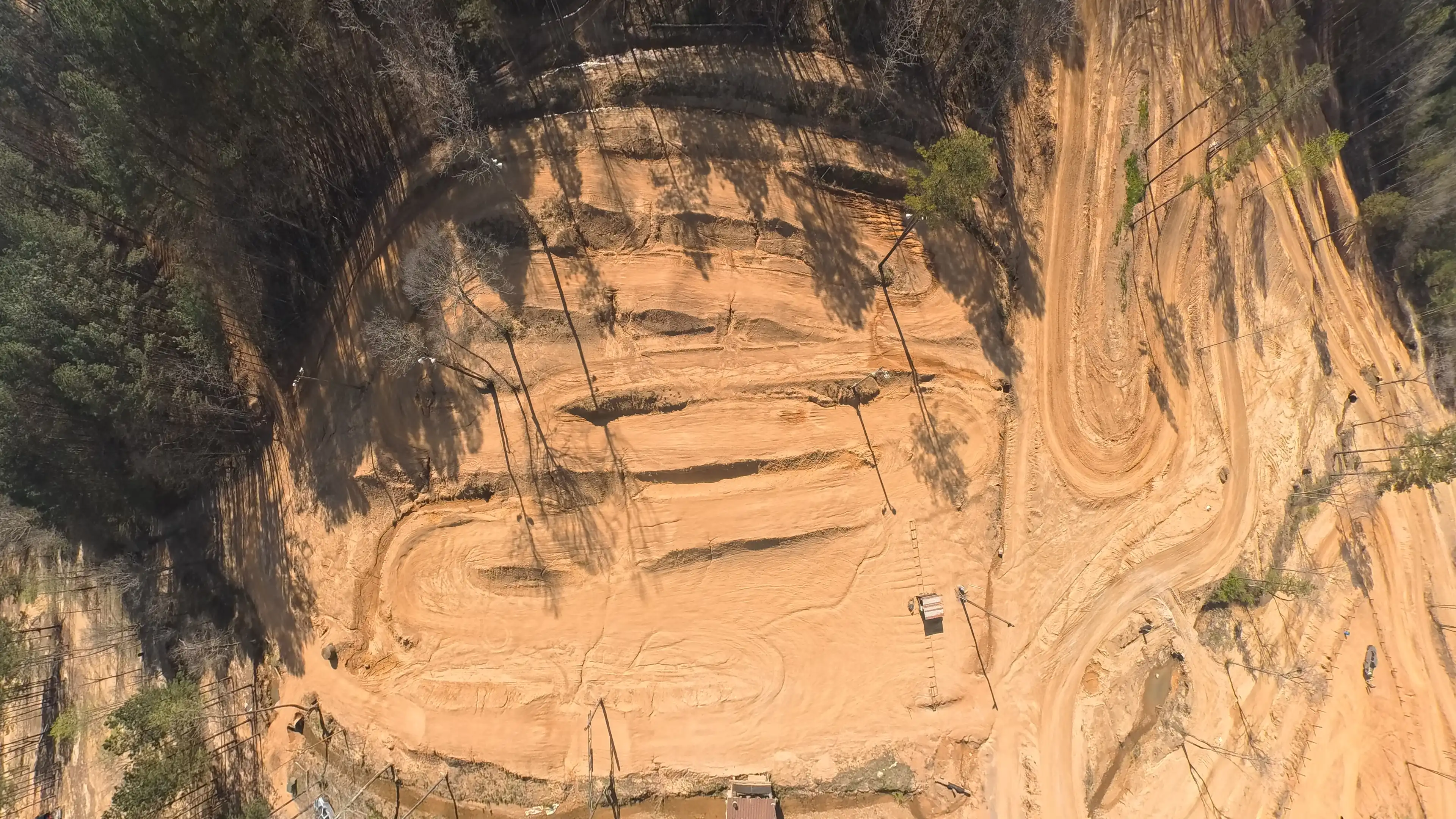 Aerial view of a dirt motocross track with jumps, turns, and surrounding trees casting long shadows.