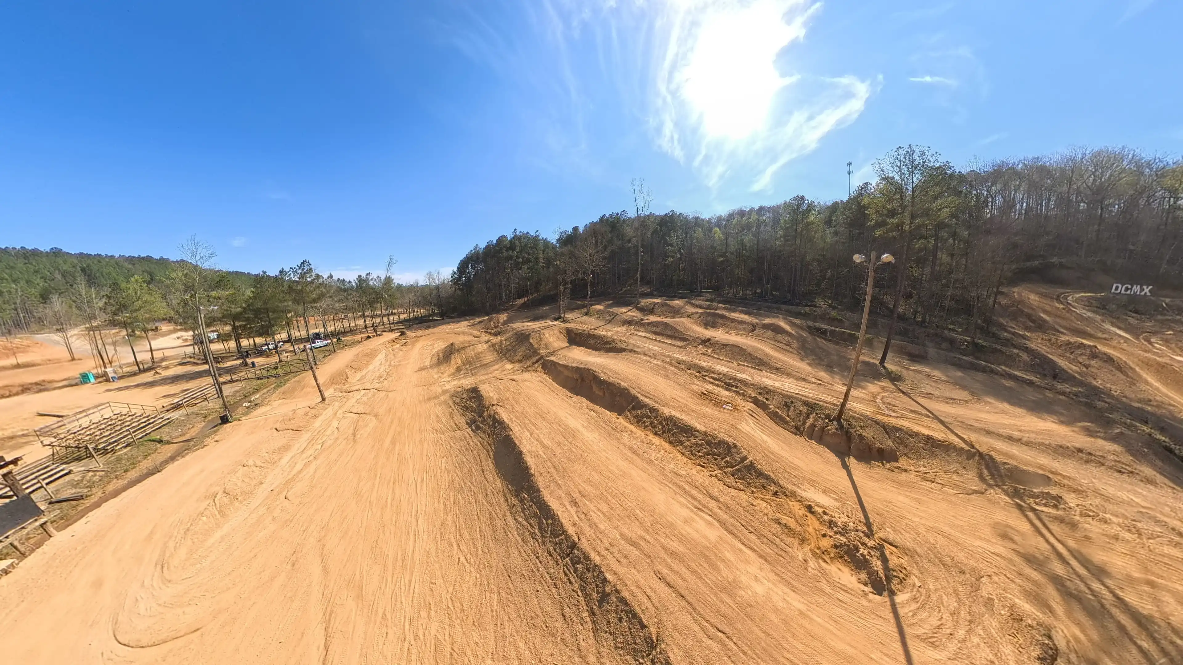 Wide view of a dirt motocross track with jumps and turns surrounded by trees under a clear sky.