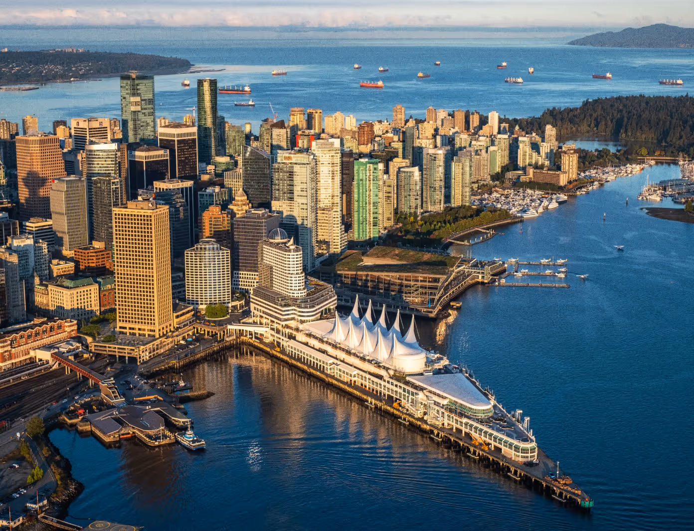 Aerial view of Vancouver waterfront with downtown skyscrapers, Canada Place sails, and cargo ships on the water.