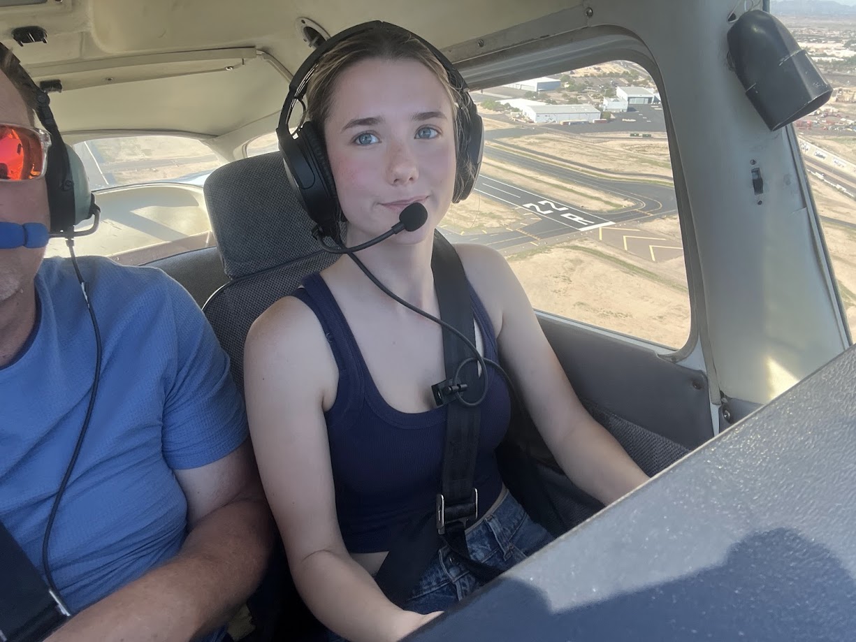 Young woman wearing a headset and seatbelt inside a small airplane cockpit with a runway visible outside the window.