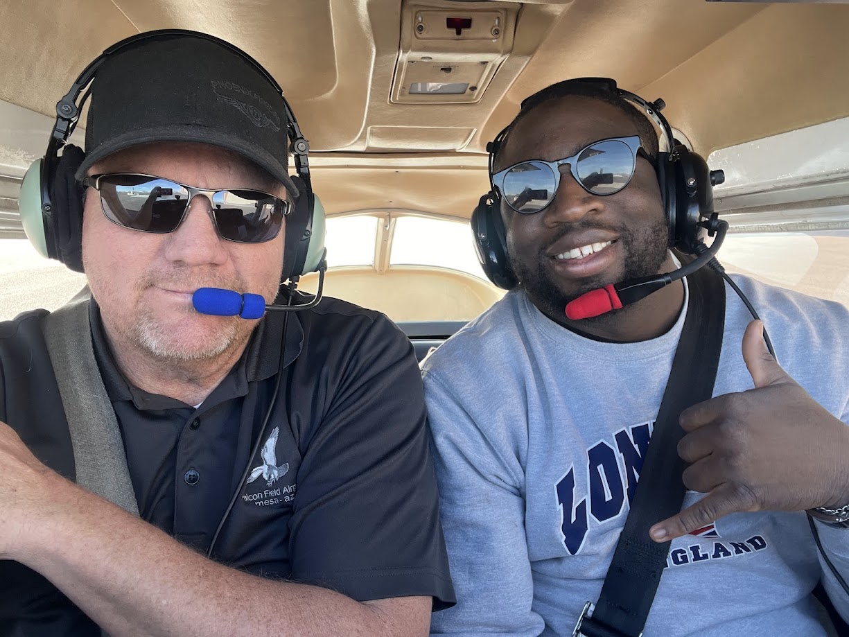 Two men wearing headsets with microphones and sunglasses sit inside a small aircraft, the man on the right gestures with a shaka hand sign.