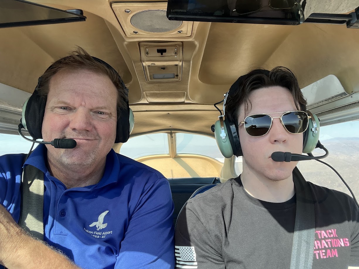 Two men wearing headsets sitting inside a small airplane cockpit, one in a blue polo and the other in a black t-shirt with sunglasses.