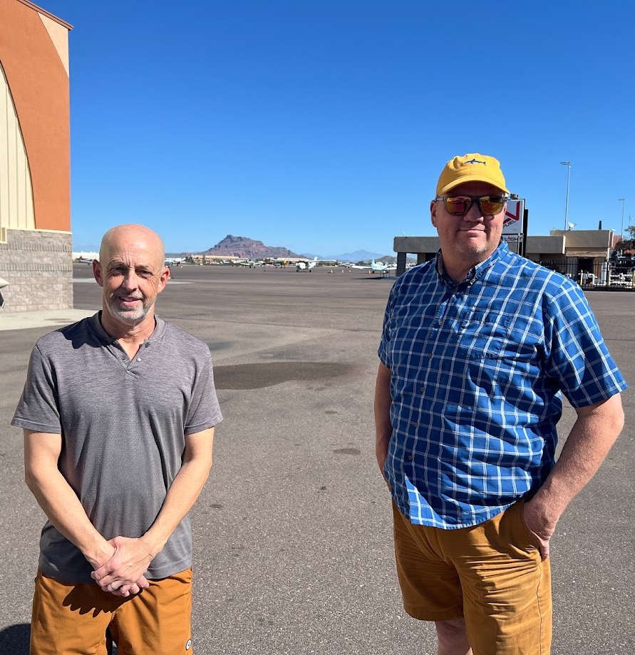 Two men standing on an airfield with a clear blue sky and small airplanes in the background.