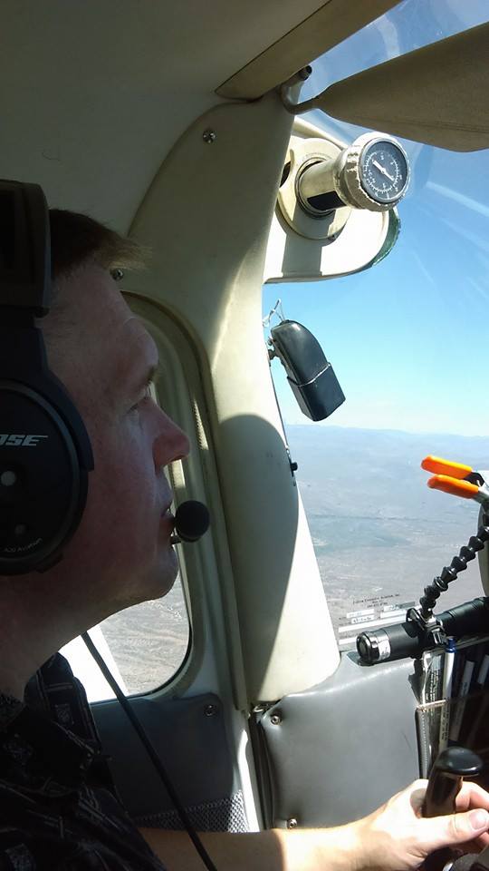 Pilot wearing headset and microphone, gripping the control yoke inside a small aircraft cockpit with a view of the landscape outside.