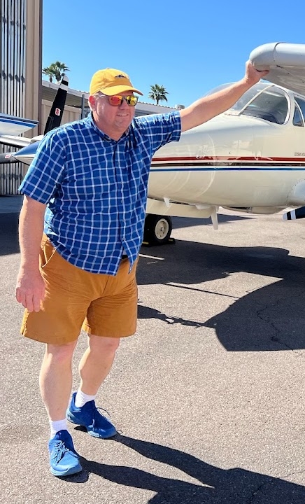 Man wearing a yellow cap, sunglasses, blue checkered shirt, and orange shorts standing beside a small airplane on a sunny day.