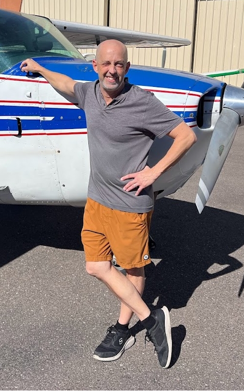 Man in a gray polo shirt and orange shorts leaning against a small white and blue airplane, standing on tarmac under sunlight.