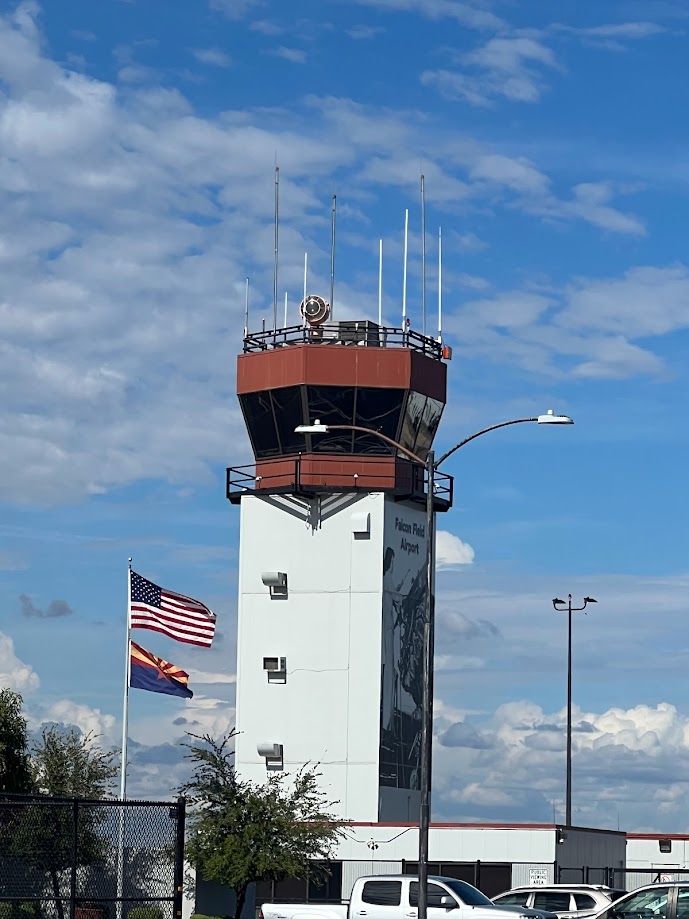 Control tower at Falcon Field Airport with American and Arizona state flags flying, clear blue sky with scattered clouds.