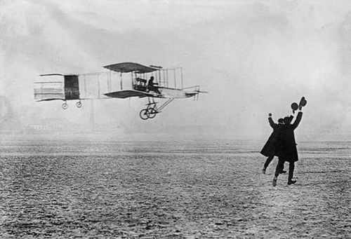 Historic black-and-white photo of an early Wright brothers' biplane in flight with two men jumping and cheering nearby on a barren field.
