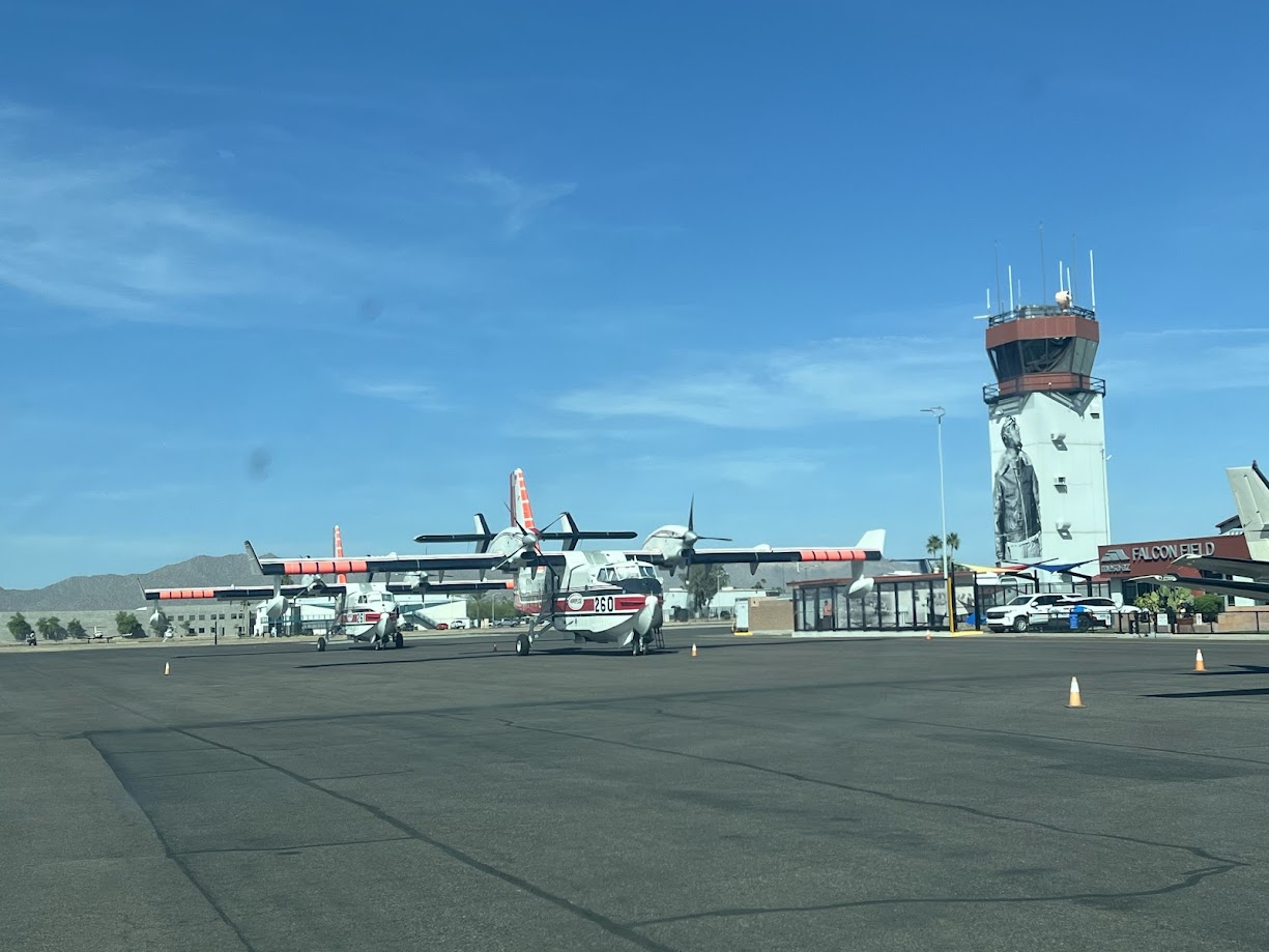 Firefighting aircraft parked on an airport tarmac with control tower and Falcon Field sign in the background.