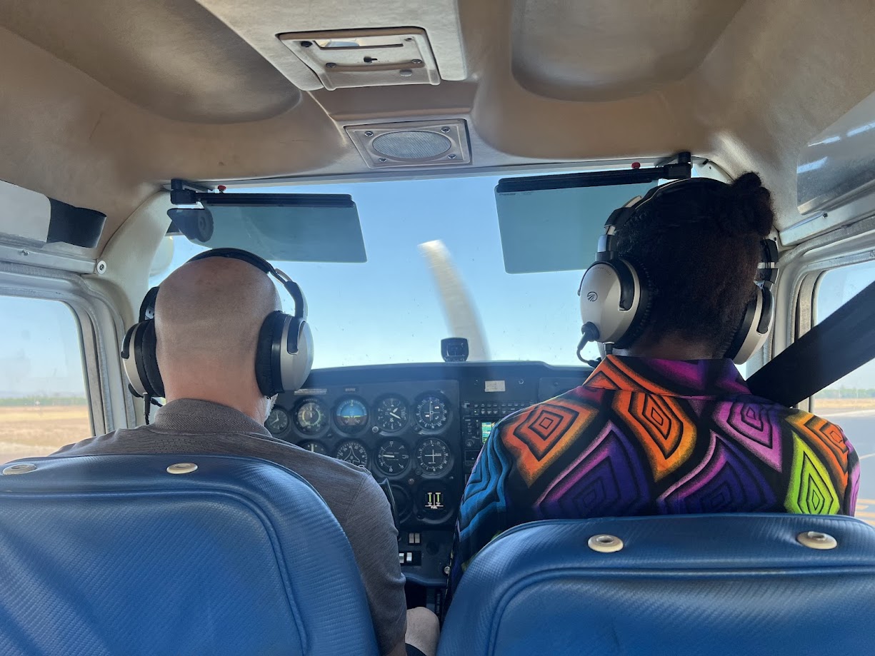 Two pilots wearing headsets seated in a small airplane cockpit facing the instrument panel and windshield.