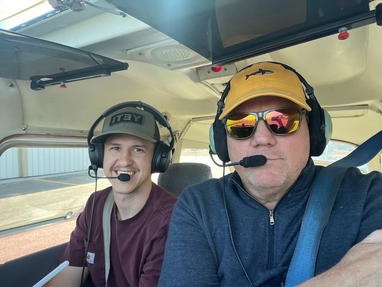 Two men inside a small airplane cockpit wearing headsets; the man on the right wears sunglasses and a yellow cap with a shark logo.