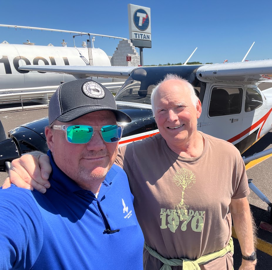 Two smiling men standing in front of a small airplane on a sunny day near a Titan sign.