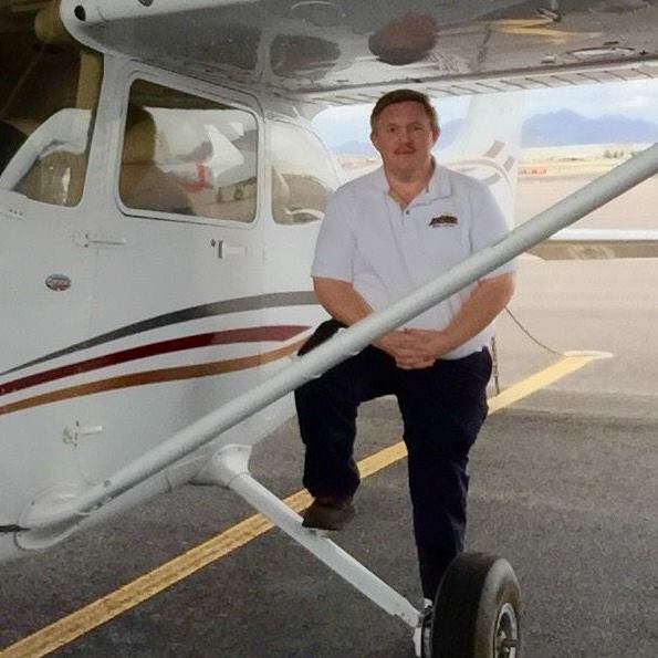 Man with a mustache and white polo shirt standing with one foot on the front wheel of a small white airplane on a runway.