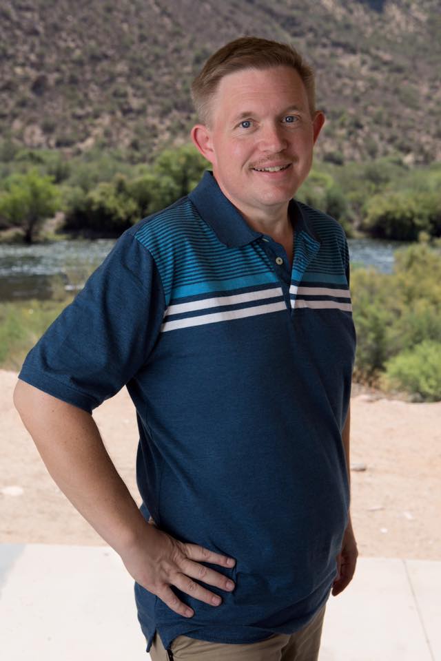 Smiling man with short light brown hair wearing a blue striped polo shirt standing outdoors near a river and trees.
