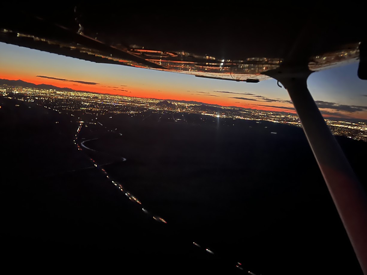 View of a city at dusk from a small airplane, showing the wing, city lights, and a colorful sunset horizon.