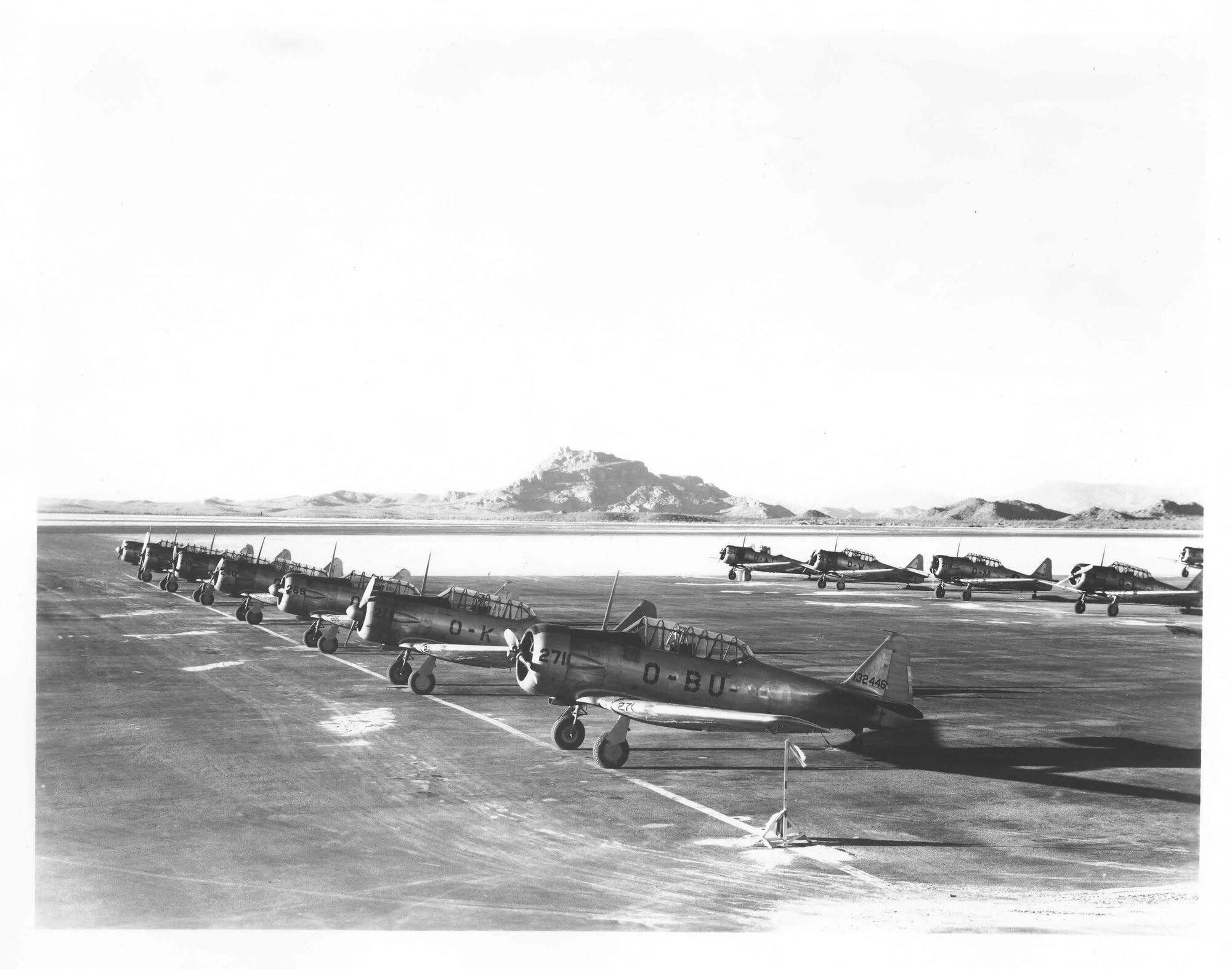 Row of vintage military training aircraft lined up on a runway with mountains in the background.