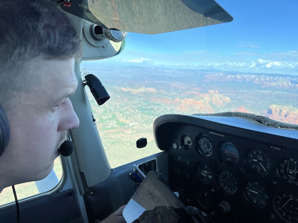 Pilot wearing a headset looking at the instrument panel inside a small airplane cockpit with a landscape view through the window.
