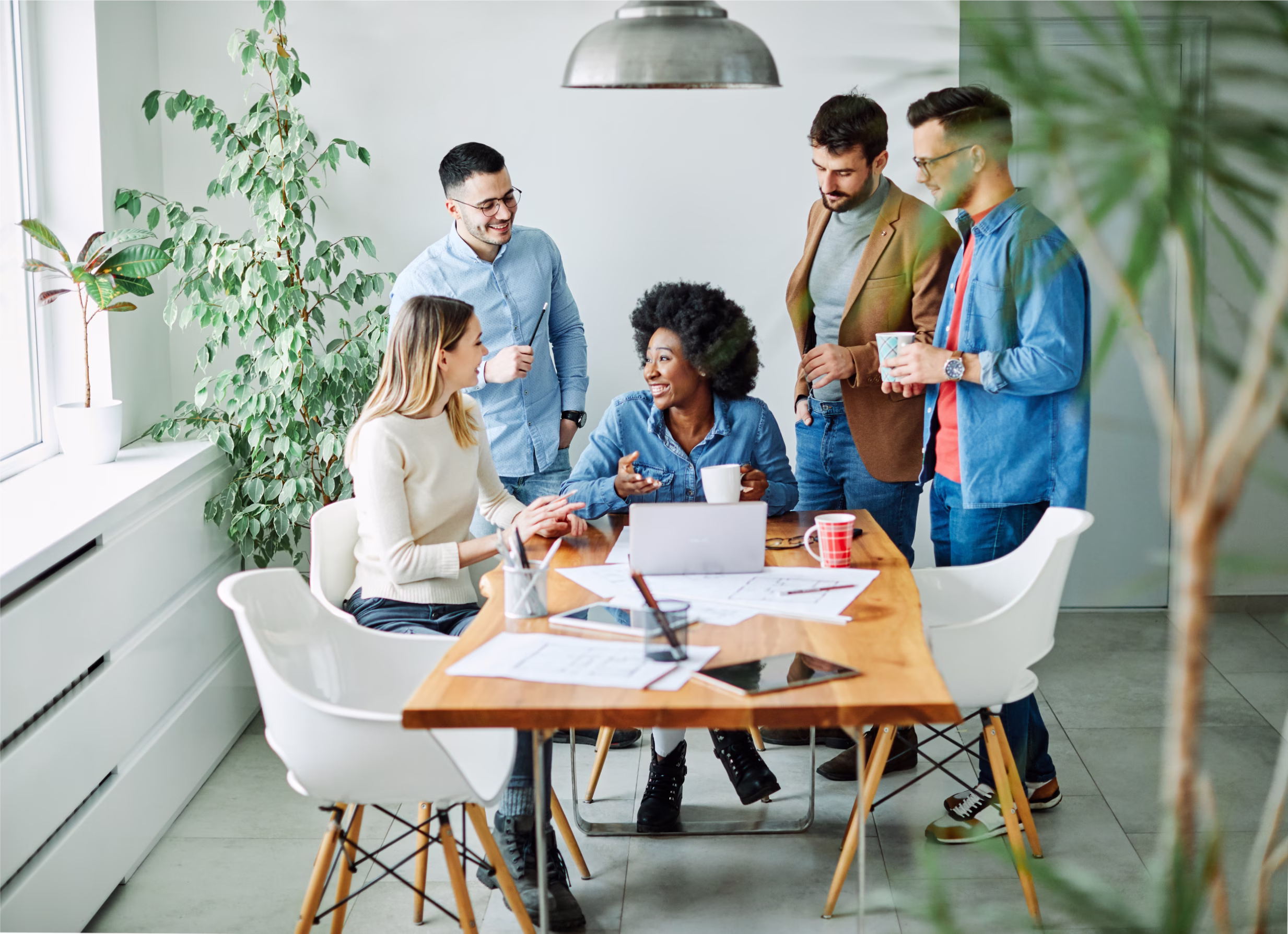 Group of five diverse coworkers smiling and discussing around a wooden table with papers and a laptop in a bright office.