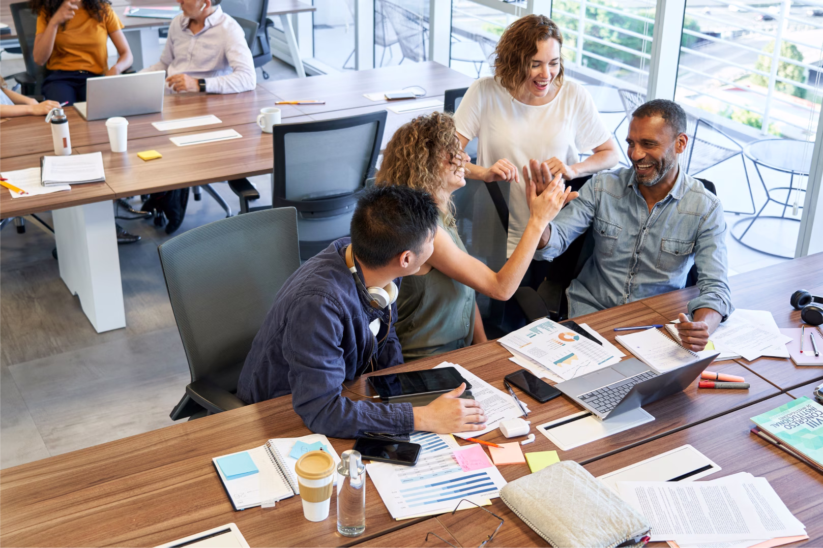 A group of diverse coworkers engaged in a meeting, with three of them giving a high-five around a wooden table covered with laptops, papers, and office supplies.