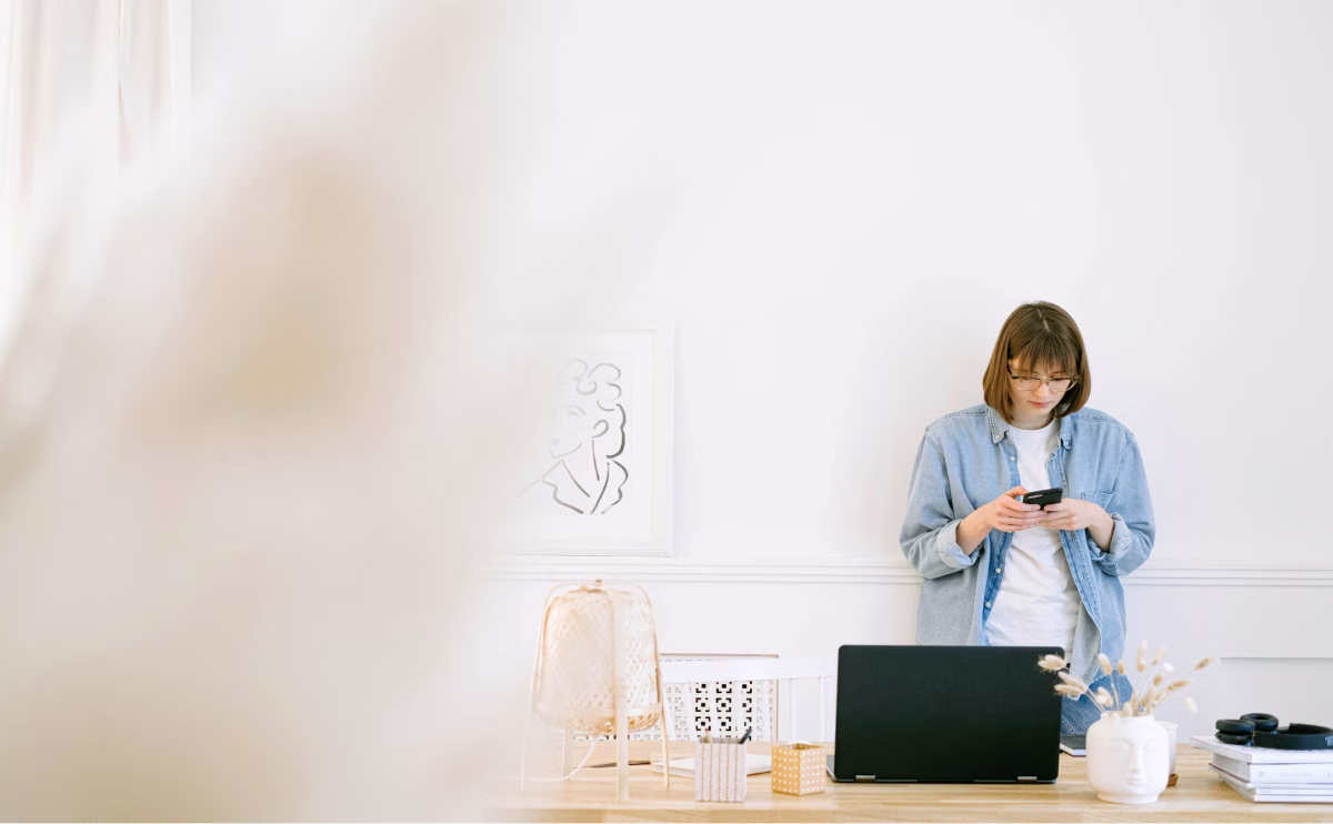 Woman in glasses and denim jacket looking at her smartphone while standing behind a desk with a closed laptop and decorative items in a bright white room.