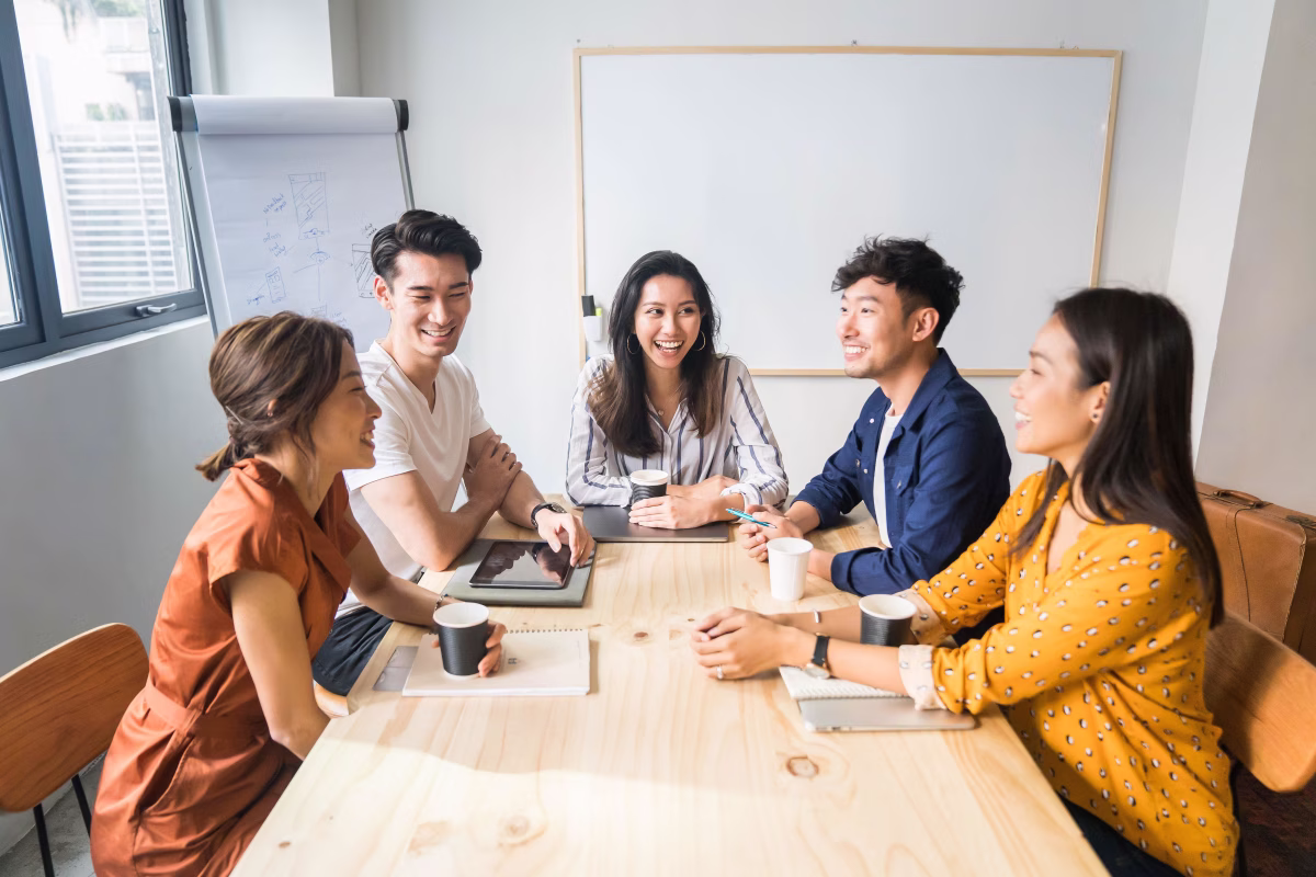 Five young professionals smiling and chatting around a wooden table in a bright meeting room with a whiteboard and flipchart.