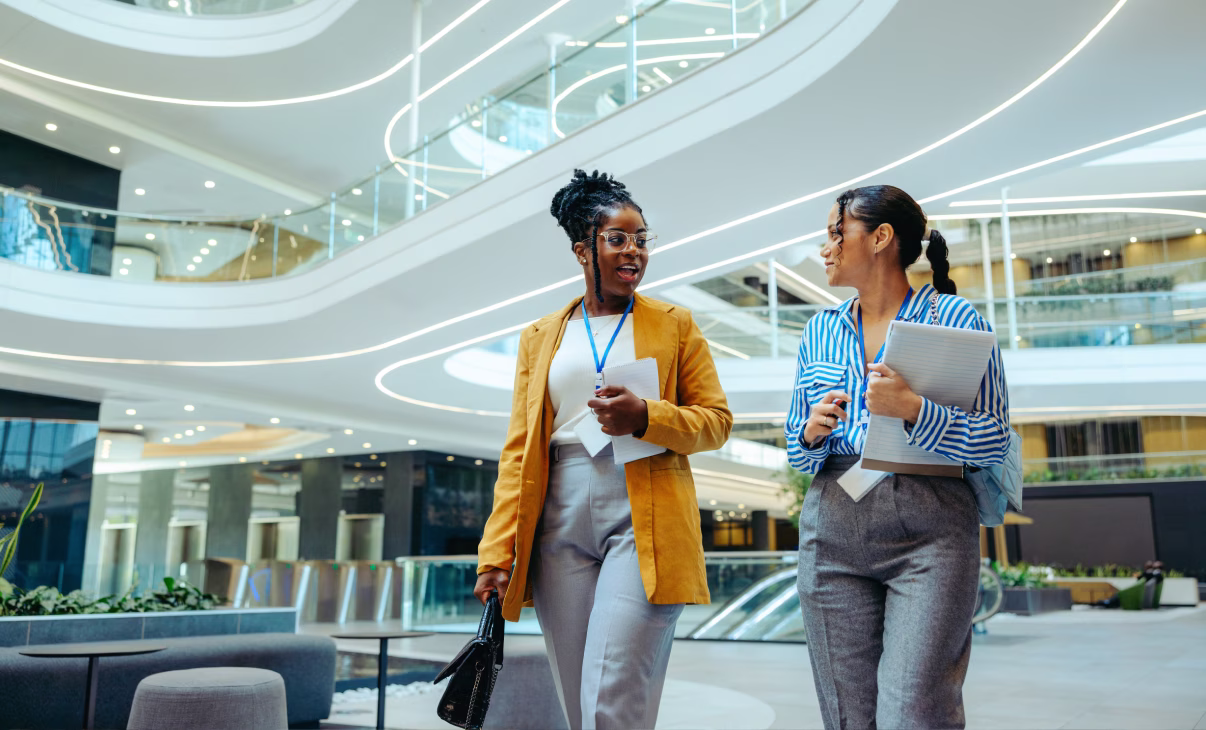 Two professional women walking and talking in a modern office lobby with curved lighting and glass railings.