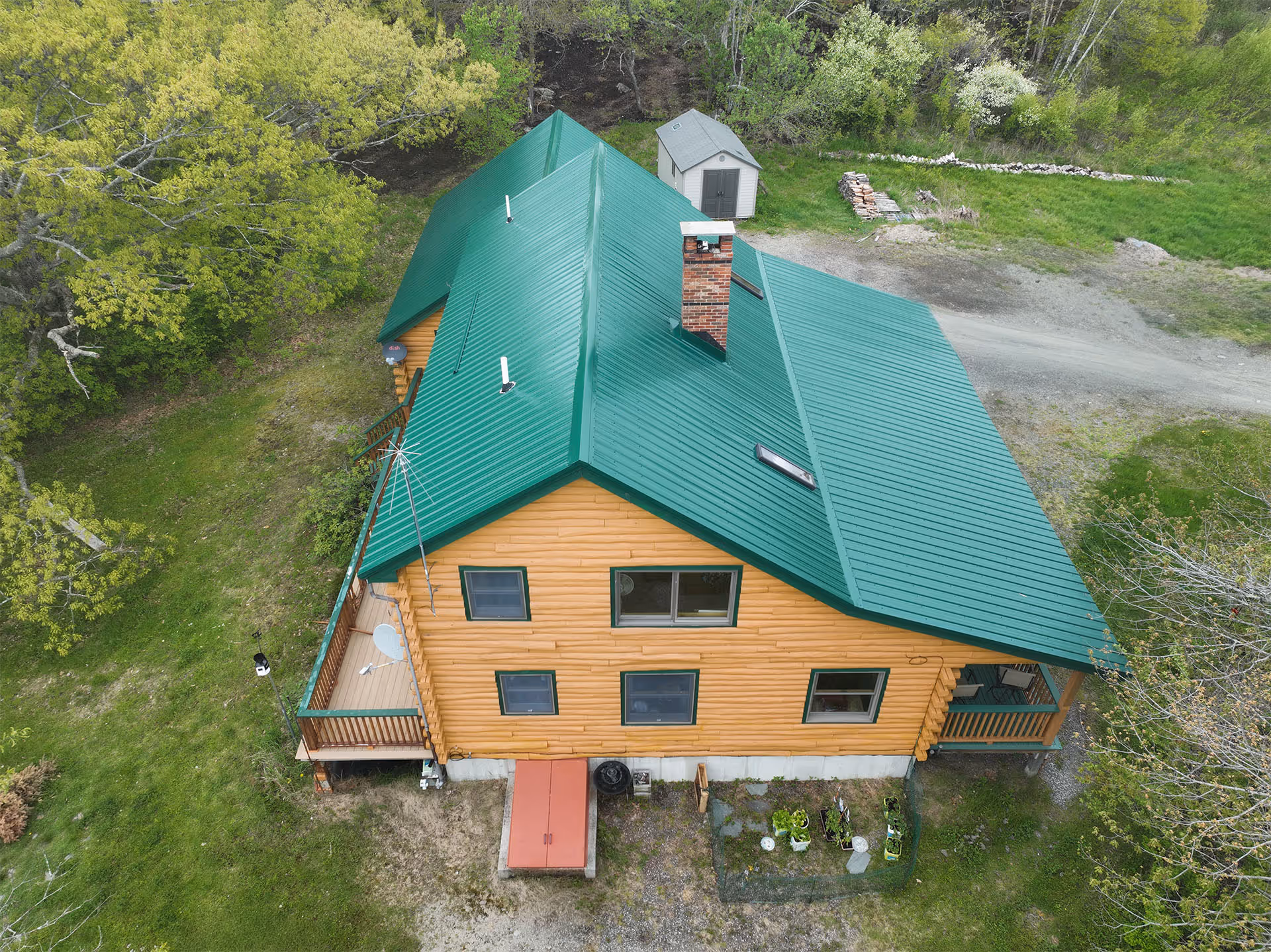 Aerial view of a log cabin with a green screw down steel roof surrounded by grass and trees.