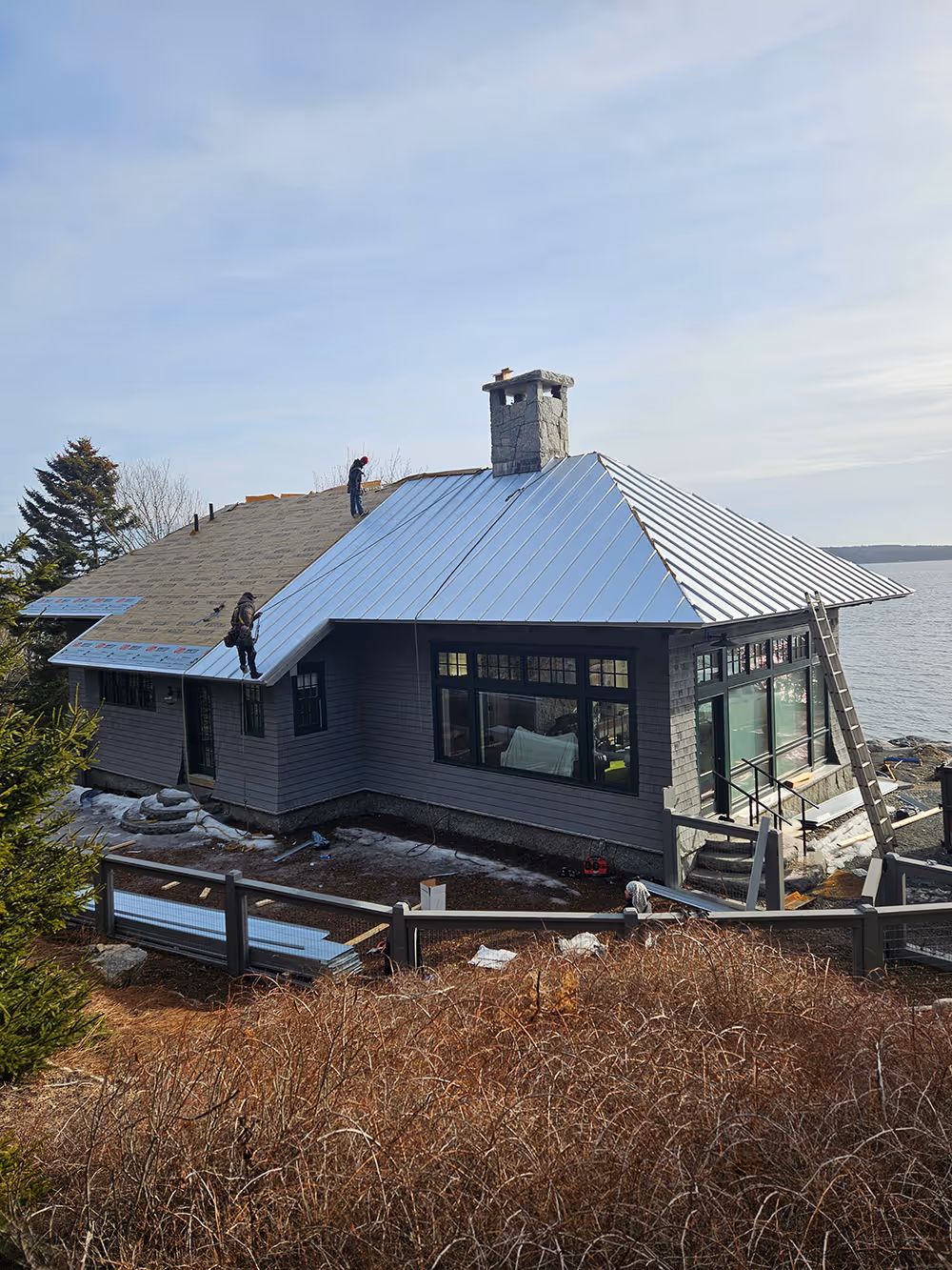 House with a partially installed metal roof by the water, with two workers on the roof and a stone chimney.