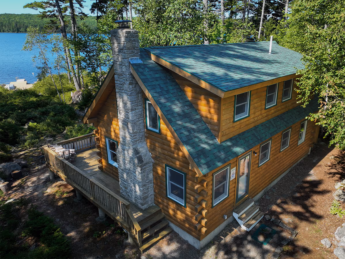 Log cabin with green shingle roofing beside a lake surrounded by trees.