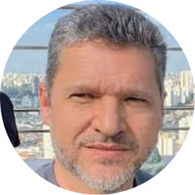 Retrato de homem adulto com cabelo curto grisalho e barba por fazer, ao ar livre com cidade ao fundo.